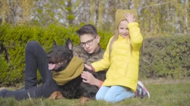 Gözlüklü genç beyaz çocuk ve sarı ceketli kız kardeşi yeşil çimlerin üzerinde, hardal eşarplı güzel doberman ile oturuyor. Neşeli kız ve erkek kardeşi dışarıda köpekleriyle vakit geçiriyorlar.