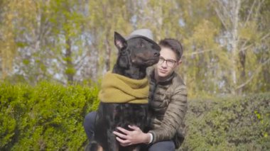 Beyaz çocuk, siyah zarif dobermana hardal eşarplı gri şapka takıyor. Genç adam yeşil çimlerde güzel bir köpekle oturuyor. Adam güz gününü hayvan arkadaşıyla dışarıda geçiriyor.