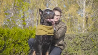 Gri şapkalı, hardal eşarplı ve güneş gözlüklü güzel siyah doberman yeşil çimlerde oturan beyaz çocuğun yanında oturuyor. Gözlüklü genç adam güz gününü hayvan arkadaşıyla dışarıda geçiriyor.
