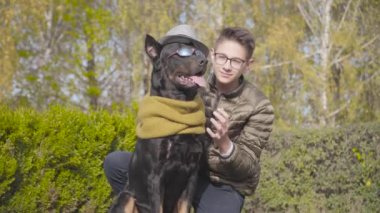 Gri şapkalı, hardal eşarplı ve güneş gözlüklü güzel siyah bir doberman ile yeşil çimlerde oturan Kafkas genci. Genç adam saçını düzeltiyor, kameraya bakıyor ve gülümsüyor.