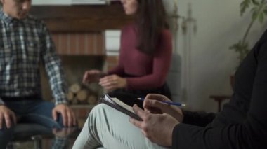 Arka planda eşine bağıran bulanık bir genç kadın. Beyaz erkek eller ön planda yazılar yazıyor. Profesyonel psikolog hastalarının konuşmalarını beklerken not alıyor..