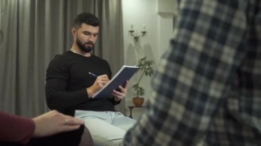 Çok profesyonel bir erkek psikoloğun portresi not alıyor ve sorular soruyor. Genç bir çift ya da aile ön planda el ele tutuşuyor. Çift terapisinin başlangıcı..