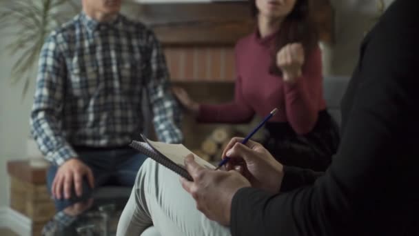 Close-up van psychologen handen die aantekeningen maken op de voorgrond. Wazige blanke vrouw die praat met haar echtgenoot en met de dokter op de achtergrond. Professionele psycholoog luisterend naar zijn patiënten. — Stockvideo