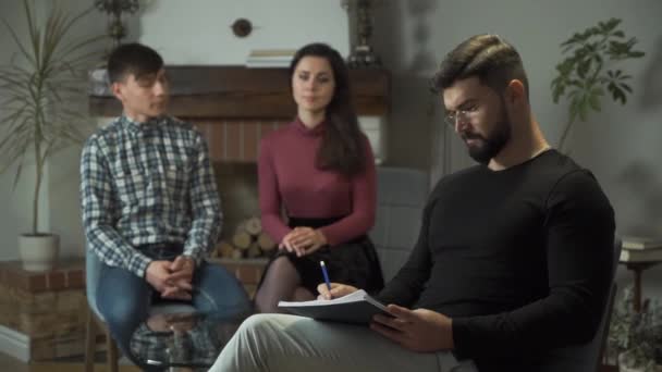 Serious Caucasian bearded man taking notes at the foreground as blurred couple taking each others hands at the background. Young handsome psychologist looking up at camera and smiling. — Stock Video