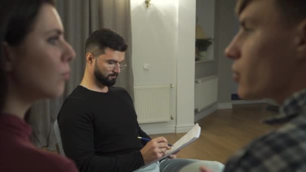 Close-up of young Caucasian man and woman talking as bearded psychologist looking at them with interest from the background. Professional psychologist at couples therapy session. Family problems. — Stock Video
