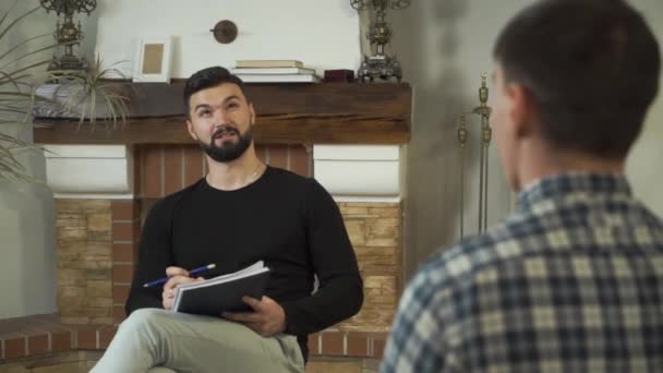 Portret van een aardige blanke man met papieren, pratend, glimlachend. Professionele mannelijke psycholoog vertelt verhaal aan patiënt die tegenover hem zit. Individuele therapie. Camera beweegt van links naar rechts. — Stockvideo