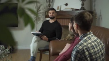 Pozitif beyaz psikolog arka planda oturuyor ve hastalarına gülümserken bakıyor. Mutlu aile, psikolojik sorunlarını profesyonel yardımla çözüyor. Başarılı terapi..