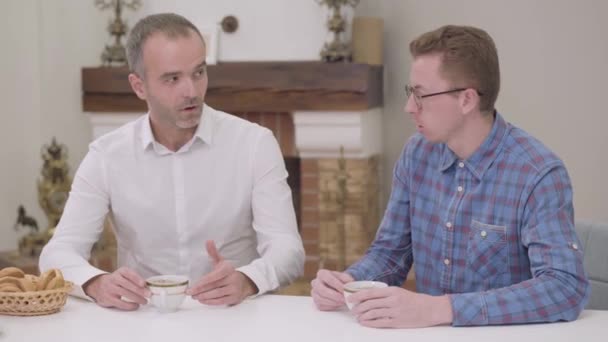 Junger kaukasischer Junge mit Brille im Gespräch mit einem älteren Mann, der am Tisch sitzt. Schwiegervater mit sarkastischem Gesicht. schwierige Beziehung zwischen Verwandten. Familientreffen, Treffen mit den Eltern, — Stockvideo