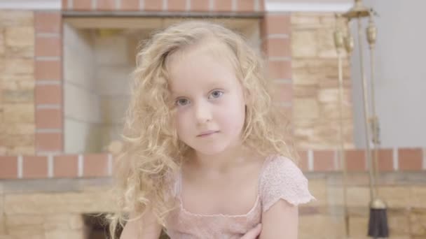 Портрет милой белокурой белокурой девушки с вьющимися волосами, смотрящей в камеру. Красивый ребенок позирует на фоне камина дома . — стоковое видео