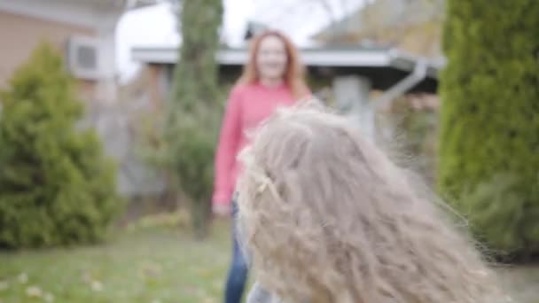 Glada kaukasiska flicka med blont lockigt hår springer runt sin leende mor i huset gården. Glad vuxen rödhårig kvinna njuter av fritid med sin dotter. Familjen vilar tillsammans utomhus. — Stockvideo