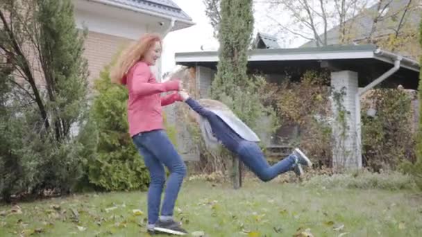 Young Caucasian redhead mother spinning daughter in the house yard. Happy little blond girl with curly hair and redhead adult woman enjoying free time together. Cheerful family having fun at weekends. — Stock Video