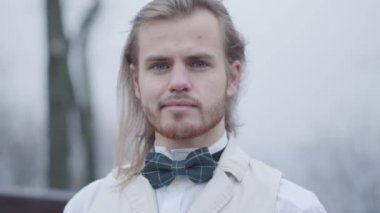 Beyaz gömlekli, ekose papyonlu beyaz bir adamın kameraya bakıp gülümsediği yakın plan yüzü. Sonbahar parkında poz veren eski tarz asil bir adam..