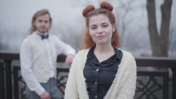 Primo piano di bella donna caucasica con doppi panini sorridenti sul ponte. Old fashioned ragazza in posa nel parco autunnale come il suo ragazzo elegante in gilet e papillon in piedi sullo sfondo . — Video Stock