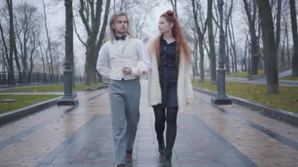 Portret van een jong Kaukasisch koppel dat in het najaarspark wandelt en praat. Gelukkige vriend en vriendin daten buiten. Man in retro stijl kleding en roodharige vrouw wandelen langs de steeg. — Stockvideo