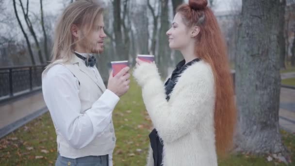 Vista lateral de la joven pareja caucásica tomando té o café en el parque de otoño y hablando. Novio y novia en ropa de estilo retro citas al aire libre . — Vídeos de Stock