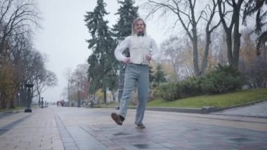 Şehir parkında dans eden zarif beyaz gömlekli, yelekli ve ekose papyonlu neşeli beyaz çocuk. Komik beyaz adam, güz gününde dışarıda eğleniyor..