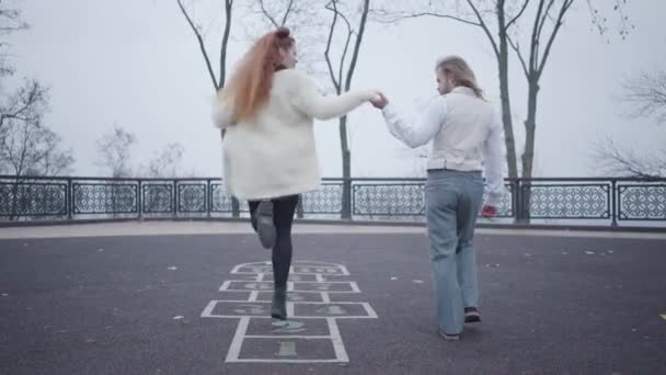 Jeune homme caucasien en vêtements élégants tenant sa main de copines alors qu'elle saute sur le jeu de marelle dessiné sur l'asphalte. Couple millénaire insouciant se reposant à l'extérieur . — Video