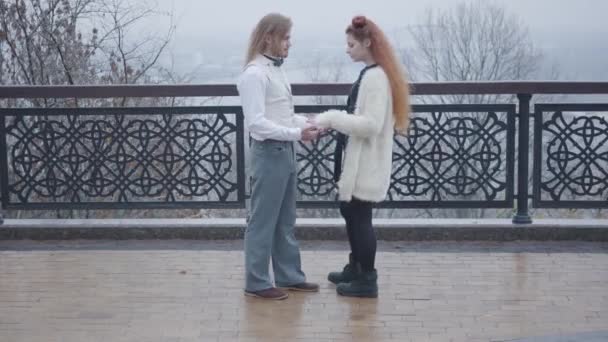 白人年轻人牵着他女朋友的手聊天的侧视图。 在秋天的公园里,一对快乐的夫妇面对面地站在一起. 旧式的男人和红头发女人，双包在户外休息. — 图库视频影像