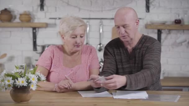 Portrét staršího bělošského páru, který se zabývá účty. Plešatý muž a blondýnka sedí u stolu a povídají si. Starší manželé vyrovnají své příjmy a výdaje. Financiály, důchodci, výplaty. — Stock video