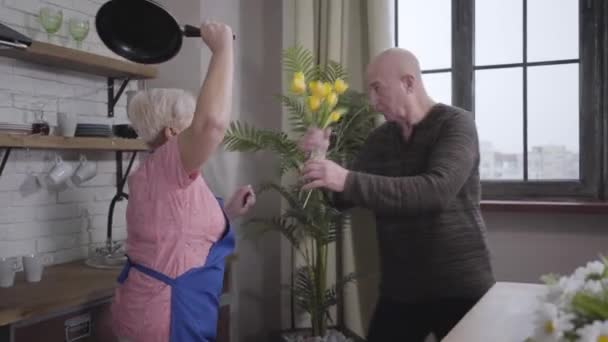 Eine ältere kaukasische blonde Frau versucht, ihren Mann mit der Pfanne zu schlagen. Reifer glatzköpfiger Mann, der sich mit einem Strauß gelber Tulpen verteidigt. Ehefrau, die ihren schuldigen Ehepartner erzieht. Rentner haben Spaß. — Stockvideo