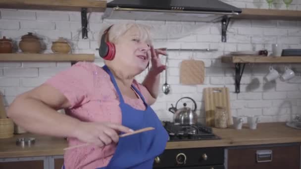 Primer plano de la mujer blanca mayor positiva bailando y haciendo gestos emocionales. Una anciana graciosa con auriculares cantando y bailando en la cocina. Maduro jubilado alegre disfrutando de la vida después de la jubilación . — Vídeo de stock