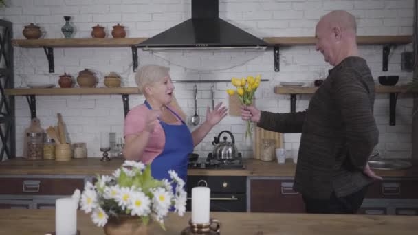 Vrolijke volwassen blanke kale man geeft boeket gele tulpen aan zijn schattige vrouw. Blond blank vrouwtje in roze T-shirt verrast tijdens het koken in de keuken. Romantiek, eeuwige liefde. — Stockvideo