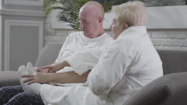 Közelkép érett fehér férfiról, aki a kanapén ülve masszírozza a feleségek lábát. A férj vigyáz az imádnivaló hitvesére. Boldog nyugdíjas pár otthon pihen. Örök szeretet, gondoskodás, összetartás. — Stock videók