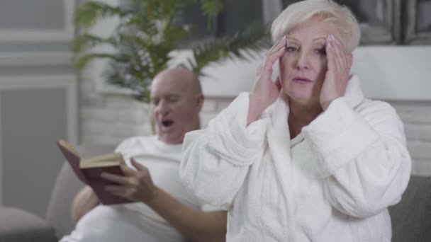 Portret van een droevige blanke vrouw met hoofdpijn zittend in witte badjas en wrijvende tempels. Haar man zit op de achtergrond en leest het boek. Veroudering, gezondheidszorg, eeuwige liefde, zorg. — Stockvideo