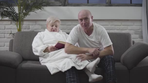 Reifer kaukasischer Mann, der mit seiner Frau auf der Couch sitzt und sich Sportspiele im Fernsehen ansieht. Frau liest Buch. Senioren ruhen sich zu Hause aus. Fan, Fußball, Ruhestand. — Stockvideo