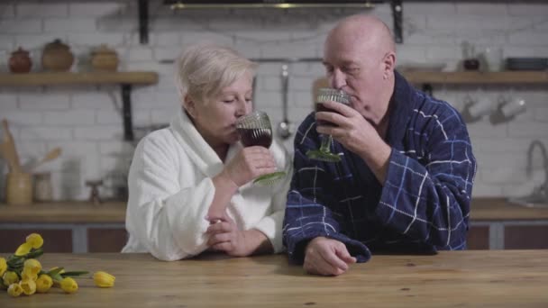 Retrato de marido y mujer caucásicos maduros felices sentados a la mesa, bebiendo vino y besándose. Los cónyuges mayores en albornoces descansan por la noche en casa. Amor eterno, felicidad, unión . — Vídeo de stock