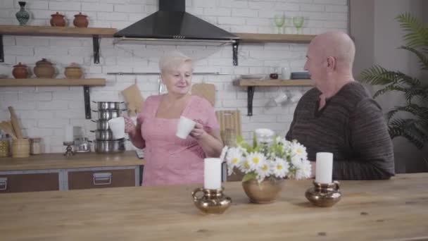 Glückliche alte kaukasische Dame bringt weiße Tasse mit Tee oder Kaffee aus der Küche und spricht mit ihrem netten Mann. lächelndes älteres Ehepaar, das sich zu Hause ausruht. ewige Liebe, Glück, Gemütlichkeit. — Stockvideo