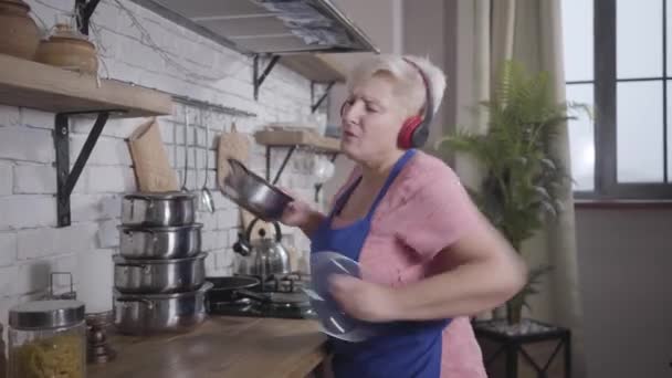Retrato de la rubia positiva abuela caucásica bailando en la cocina con cacerola. Feliz mujer sonriente en auriculares disfrutando de la cocina. Jubilado alegre . — Vídeo de stock