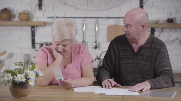 Traurige reife kaukasische Frau, die Rechnungen mit ihrem Mann beglichen. Seniorchef beruhigt seine Frau und lächelt. Ehegatten, die ihre Einkünfte und Ausgaben begleichen. Finanzen, Rentner, Auszahlungen. — Stockvideo