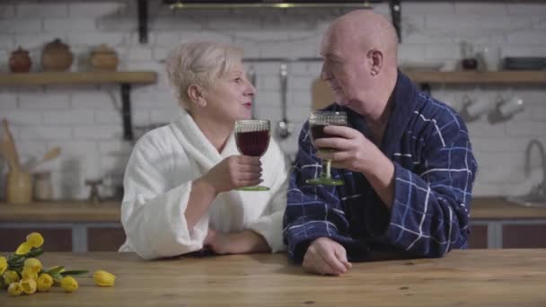 Souriant mari et femme caucasiens aînés assis à la table avec des verres à vin et de parler. Anciens conjoints en peignoirs de bain se reposant le soir à la maison. Amour éternel, bonheur, convivialité . — Video