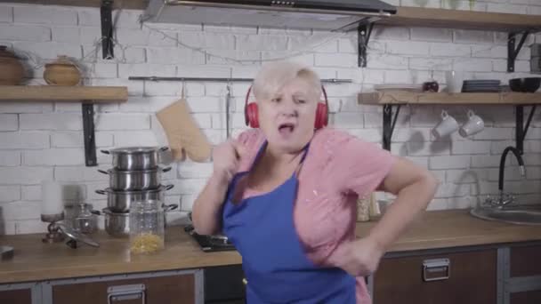 Vicces fehér öregasszony fülhallgatóban énekel és táncol a konyhában. Idős, vidám nyugdíjas, aki a nyugdíjazás után élvezi az életet. — Stock videók