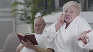 Ön planda telefonla konuşan, kahverengi gözlü, sarı saçlı, yaşlı, pozitif, beyaz bir kadın. Sinirli koca arka planda dikilip gidiyor. Emeklilik, dinlenme, ev içinde.