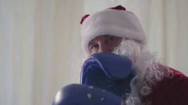 Kameraya karşı boks eldivenli sakallı Noel Baba 'nın yakın plan yüzü. Yaşlı adam tehditkar bir şekilde havayı yumrukluyor. Noel, tatil, kötü Noel Baba.
