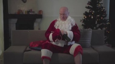 Noel Baba kılığındaki kıdemli beyaz adam kırmızı şapka takıp televizyonu açıyor. Bir şişe birayla kanepede oturan yorgun yaşlı adam..