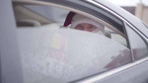 Viejo Santa Claus abriendo la ventana del coche y guiñando un ojo. Santa barbudo sentado en el coche en el asiento trasero con las cajas presentes. Navidad, vacaciones, concepto de regalos . — Vídeos de Stock