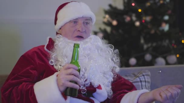 Primo piano del triste uomo caucasico in costume da Babbo Natale che mastica e beve birra. Babbo Natale cattivo seduto sul divano accanto all'albero di Natale. Alcolismo, depressione, negativismo . — Video Stock