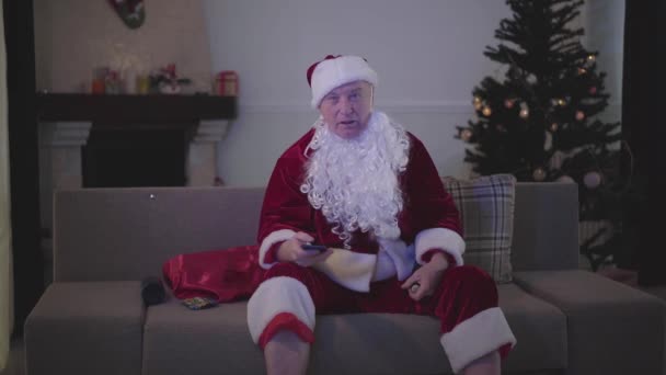 身穿圣诞老人服装、头戴假白胡子的老人坐在沙发上看电视。 成年男子在辛苦工作日后在家里休息 — 图库视频影像