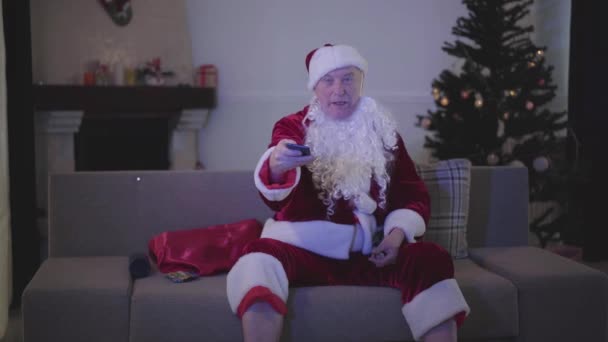 Portret starego białego alkoholika Mikołaja zmieniającego kanały i szukającego alkoholu do picia. Bad Santa Claus spoczywa na kanapie w tle choinki. — Wideo stockowe