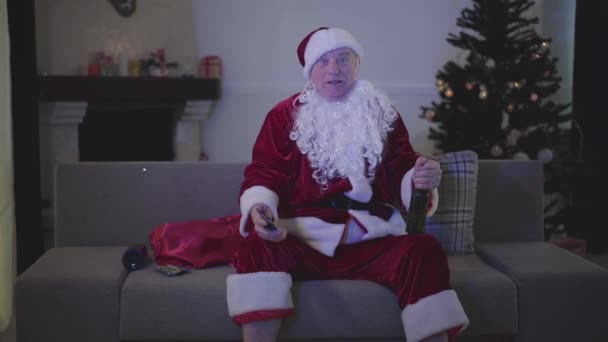 一个身穿圣诞老人服装的积极的白人男子的画像，他坐在沙发上喝啤酒，在家里看电视。 老年人看喜剧节目，辛苦了一天后在家里休息. — 图库视频影像