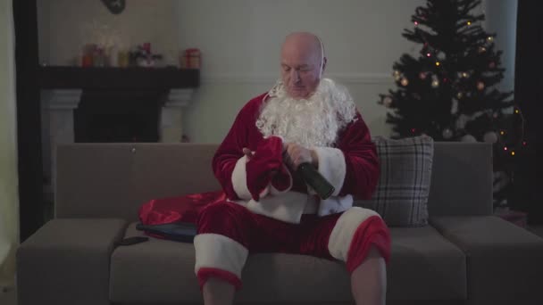 Uomo anziano caucasico vestito da Babbo Natale che si mette il cappello rosso e accende la TV. Vecchio stanco seduto sul divano con una bottiglia di birra . — Video Stock