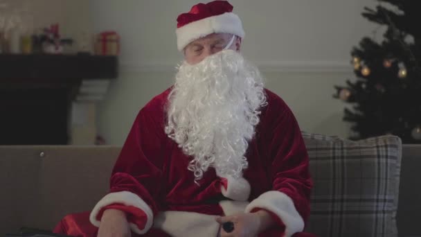 Vecchio ubriaco con la barba finta storto in faccia nel costume di Babbo Natale seduto sul divano a casa, con in mano una bottiglia vuota. Babbo Natale cattivo. Alcolismo, depressione, negativismo, solitudine — Video Stock