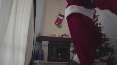 Noel Baba kostümü içinde tanınmayan bir adam yeni yıl ağacının önünde elinde hediye çantasıyla dans ediyor. Mutlu bayramlar, gelenekler, Noel kavramı. Alt görünüm