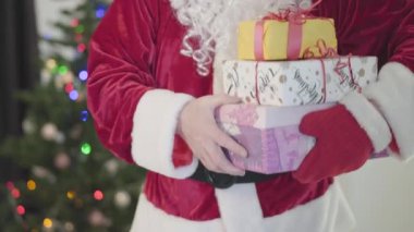 Noel Baba kostümlü yaşlı beyaz adam yılbaşı ağacının önünde kameraya hediyeler veriyor. Hediye kutuları tutan adam. Mutlu bayramlar, gelenekler, Noel kavramı.