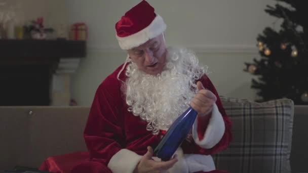 Homem branco bêbado morto em traje de Papai Noel sentado no sofá em casa segurando garrafa vazia. Homem maduro derramando as últimas gotas na mão e lambendo. Mau Pai Natal, alcoolismo . — Vídeo de Stock