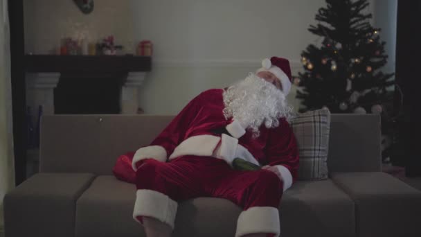Portrét dospělého bělocha v kostýmu Santa Clause spícího na gauči s lahví piva. Stařík tráví prázdniny sám doma. Špatný Santa, osamělost, deprese, závislost na alkoholu. — Stock video