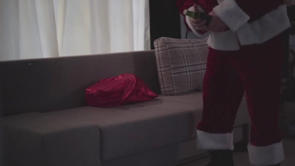 Fylld gammal man med falskt skägg i ansiktet i jultomtens dräkt sitter på soffan hemma och försöker öppna flaskan med tänderna. Dålig tomte. Alkoholism, depression, negativism, ensamhet — Stockvideo